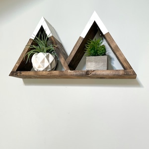 May include: A wooden shelf with a white painted top, shaped like two triangles. The shelf is mounted on a white wall and has two small potted plants on it.
