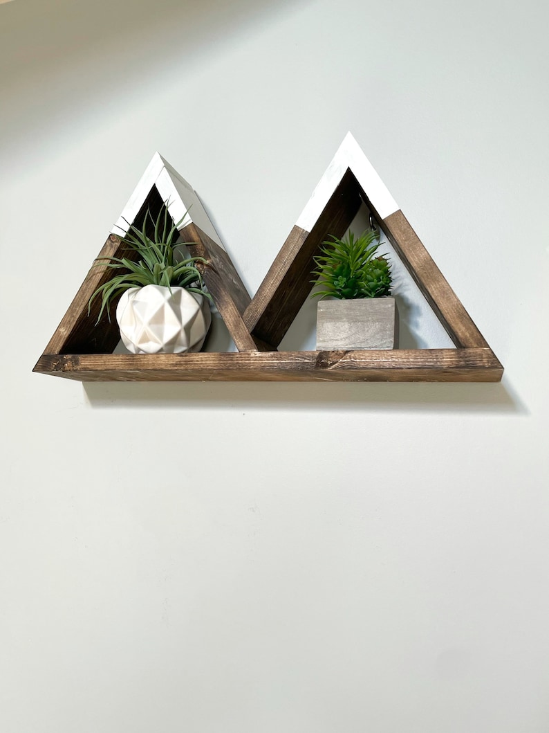 May include: A wooden shelf with a white painted top, shaped like two triangles. The shelf is mounted on a white wall and has two small potted plants on it.