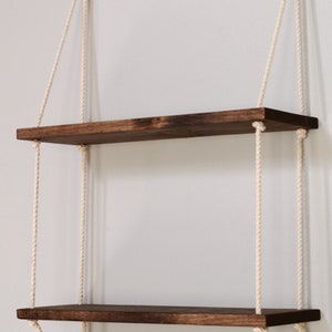 May include: Two wooden shelves hanging from the ceiling with white ropes. The shelves are stained a dark brown colour.