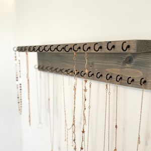 May include: A wooden jewelry organizer with multiple hooks for hanging necklaces. The organizer is painted a light gray color and has a rustic finish. The hooks are black metal and are arranged in two rows. Several gold necklaces are hanging from the hooks.