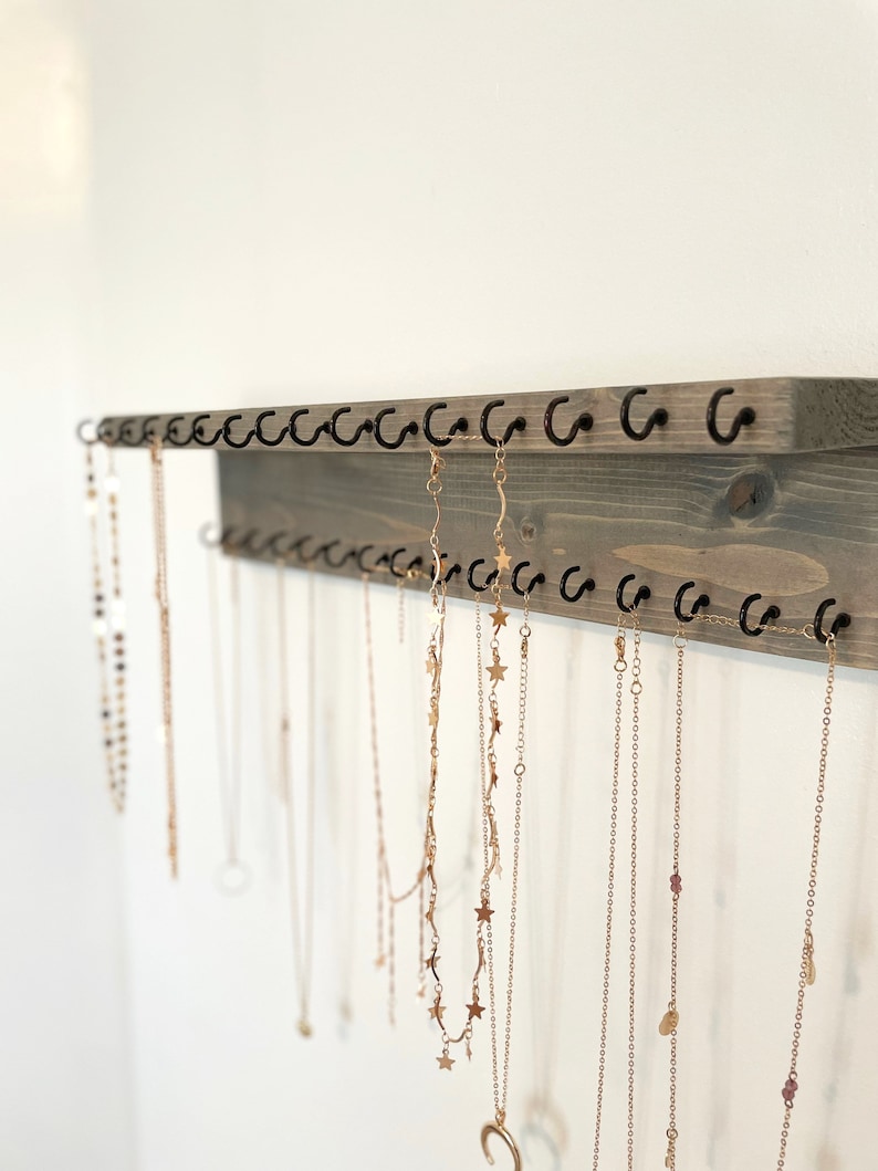 May include: A wooden jewelry organizer with multiple hooks for hanging necklaces. The organizer is painted a light gray color and has a rustic finish. The hooks are black metal and are arranged in two rows. Several gold necklaces are hanging from the hooks.