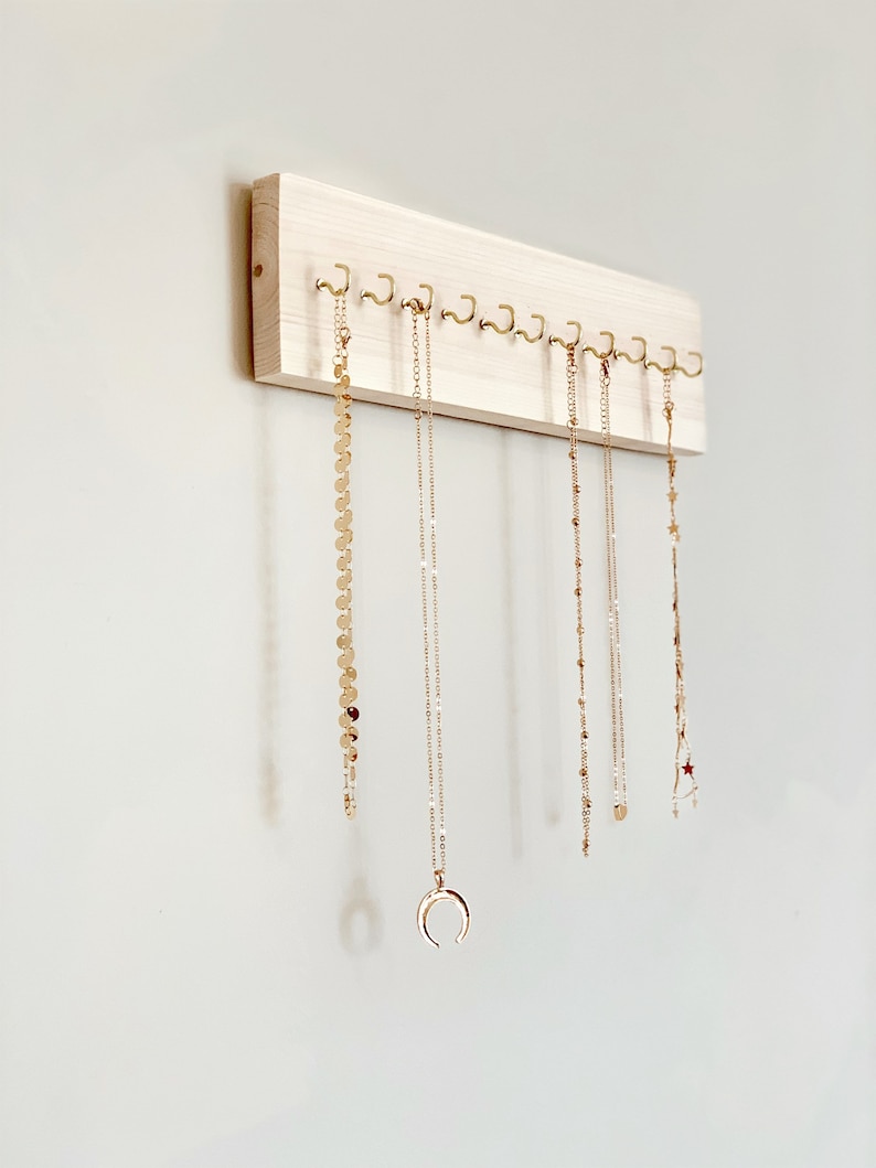 May include: A wooden jewelry organizer with gold hooks, holding several gold necklaces with different pendants. The organizer is mounted on a white wall.