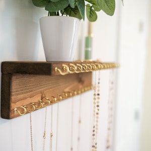 May include: A wooden shelf with gold hooks for hanging necklaces. The shelf is mounted on a white wall and has a small potted plant on top.