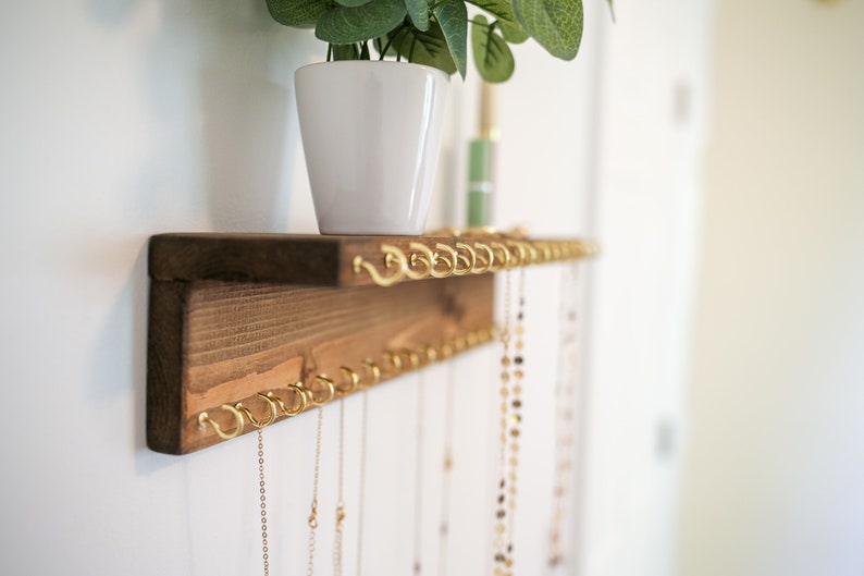 May include: A wooden shelf with gold hooks for hanging necklaces. The shelf is mounted on a white wall and has a small potted plant on top.