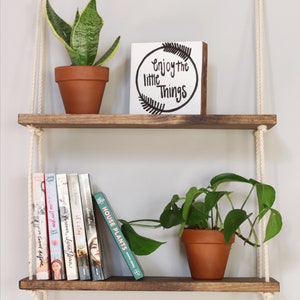 May include: Two wooden shelves suspended from the ceiling with rope. The top shelf features a potted snake plant and a white sign reading "Enjoy the little things". The bottom shelf displays a potted plant and a stack of books.
