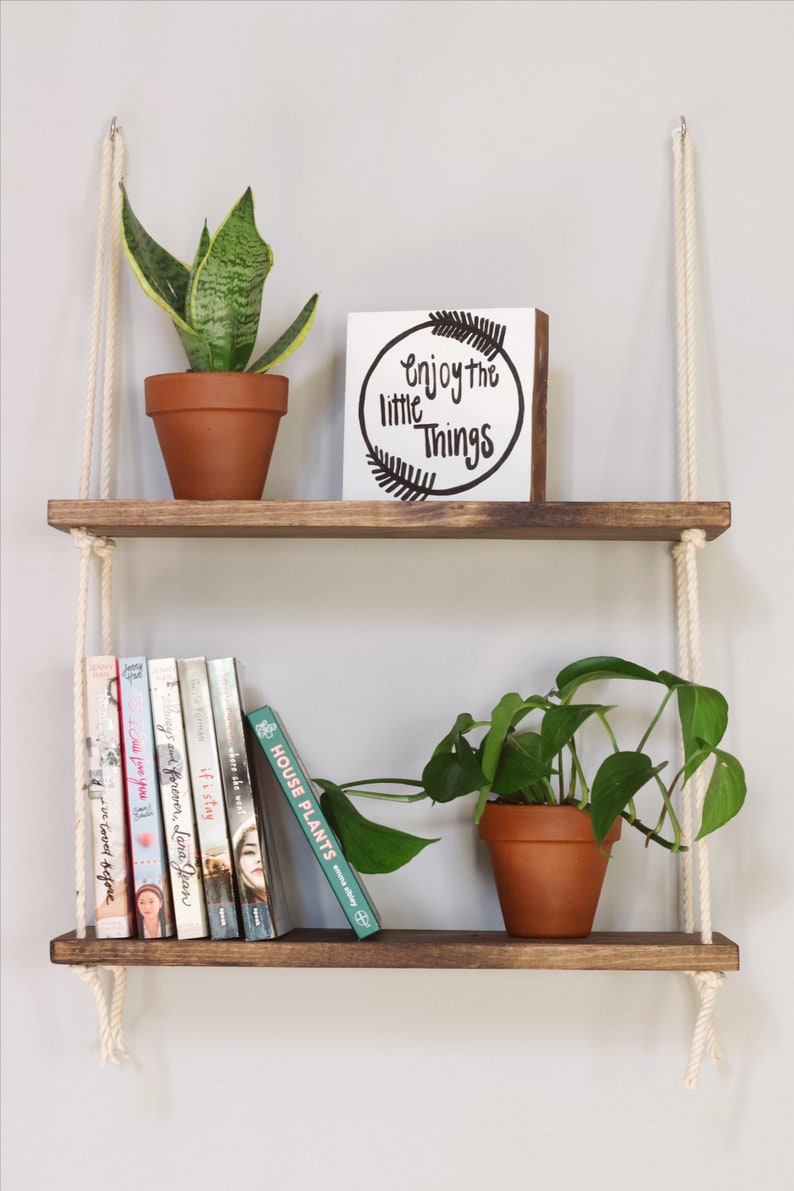 May include: Two wooden shelves suspended from the ceiling with rope. The top shelf features a potted snake plant and a white sign reading "Enjoy the little things". The bottom shelf displays a potted plant and a stack of books.