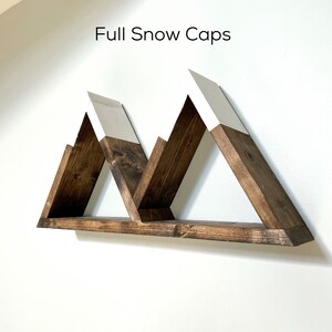 May include: A wooden shelf shaped like three mountains with white snow caps. The shelf is mounted on a white wall. The text "Full Snow Caps" is written above the shelf.