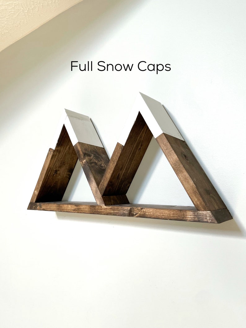 May include: A wooden shelf shaped like three mountains with white snow caps. The shelf is mounted on a white wall. The text "Full Snow Caps" is written above the shelf.