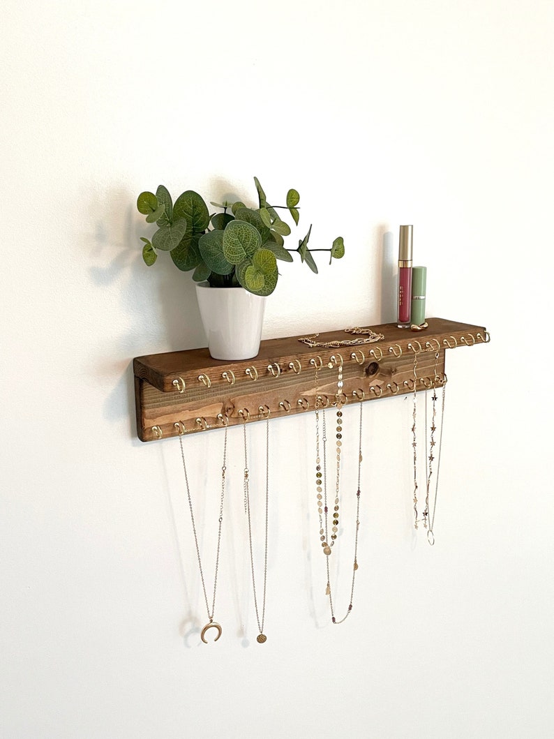 May include: Wooden jewelry organizer with a shelf and hooks for hanging necklaces. The organizer is mounted on a white wall and has a small potted plant on the shelf.