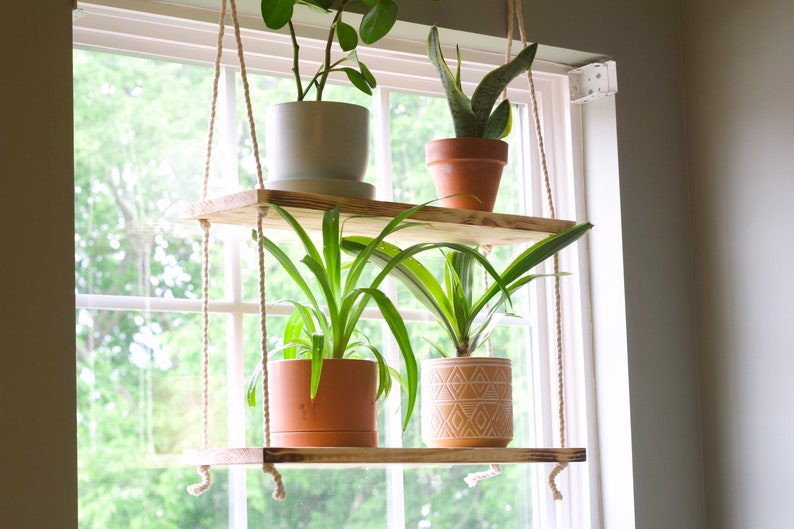 May include: Two wooden shelves suspended from the ceiling by ropes, holding potted plants. The shelves are mounted in front of a window with a view of green trees.