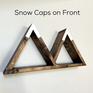 May include: A wooden shelf shaped like two mountains with white snow caps on top. The shelf is mounted on a white wall. "Snow Caps on Front" is written above the shelf.