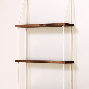 May include: Three wooden shelves with a dark brown stain, hanging from the ceiling with white ropes. The shelves are evenly spaced and are suspended from the ceiling with two ropes each.