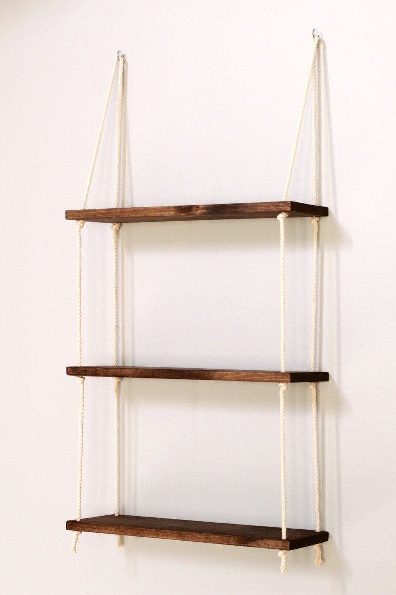 May include: Three wooden shelves with a dark brown stain, hanging from the ceiling with white ropes. The shelves are evenly spaced and are suspended from the ceiling with two ropes each.
