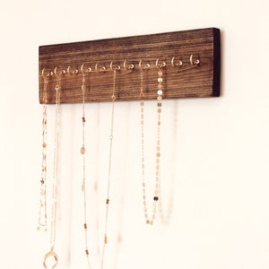 May include: A dark brown wooden jewelry organizer with gold hooks. Five gold necklaces are hanging from the hooks.