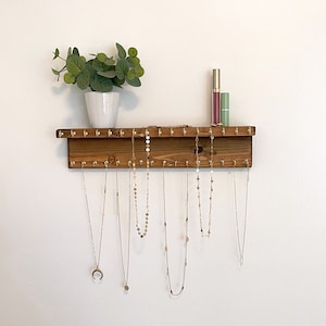 May include: Wooden jewelry organizer with gold hooks, featuring a variety of necklaces hanging from the hooks. The organizer is mounted on a white wall with a small potted plant above it.