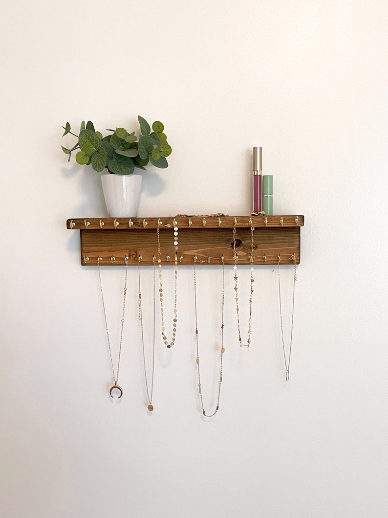 May include: Wooden jewelry organizer with gold hooks, featuring a variety of necklaces hanging from the hooks. The organizer is mounted on a white wall with a small potted plant above it.