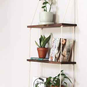 May include: Three wooden shelves with rope hangers, each shelf is decorated with plants and books. The top shelf has a green plant in a light green pot. The middle shelf has a green plant in a terracotta pot and a stack of books. The bottom shelf has a green plant in a terracotta pot and a sign that says "Enjoy the Little Things".