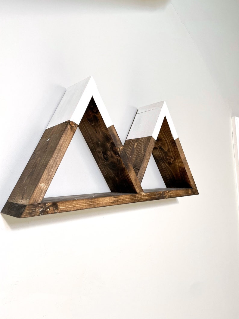 May include: A wooden shelf shaped like three mountains with white painted peaks. The shelf is mounted on a white wall.
