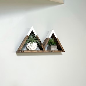 May include: A set of two triangular wooden shelves painted white on the top and brown on the bottom. The shelves are mounted on a wall and are decorated with small potted plants.