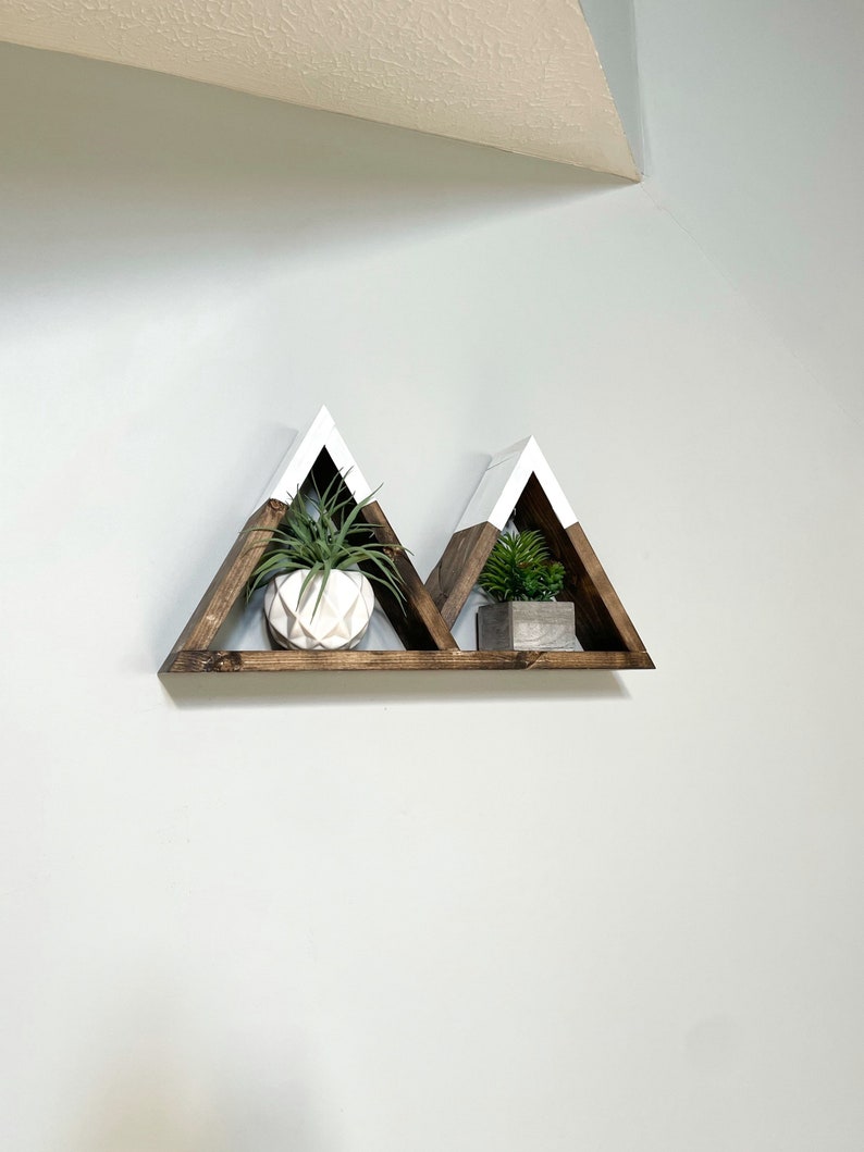 May include: A set of two triangular wooden shelves painted white on the top and brown on the bottom. The shelves are mounted on a wall and are decorated with small potted plants.