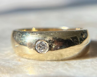 May include: A gold ring with a single, small, round diamond set in the center. The ring has a thick band and a polished finish, reflecting light. The ring is set on a light-colored surface.