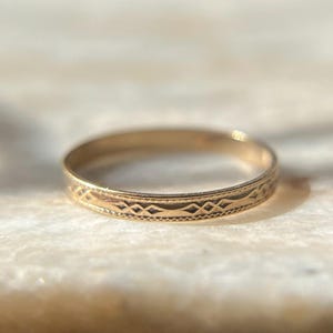 May include: A thin gold band ring featuring a delicate, repeating geometric pattern engraved along the outer surface. The design consists of small diamond shapes connected by fine lines, bordered by a subtle beaded texture on both edges.