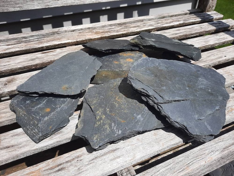 Pu&ograve; includere: Un mucchio di piastrelle di ardesia grigio scuro, alcune con un tocco di oro, disposte su una superficie di legno.