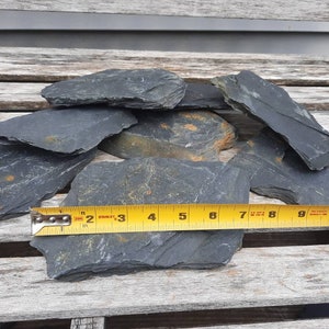 Pu&ograve; includere: Un mucchio di pezzi di roccia di ardesia grigio scuro, alcuni con una leggera sfumatura dorata, misurati con un metro a nastro giallo segnato in pollici.