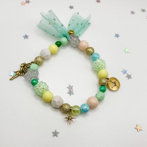 Könnte beinhalten: Ein grünes, gelbes und rosafarbenes Perlenarmband mit einem Feen-Anhänger, einem Stern-Anhänger und einem Kompass-Anhänger. Das Armband hat eine hellblaue Tüllschleife.