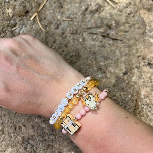 May include: Three bracelets stacked on a wrist. The top bracelet is yellow with white beads that spell "MAGIC MOMMA". The middle bracelet is pink with white beads. The bottom bracelet is yellow with a gold charm of a building.