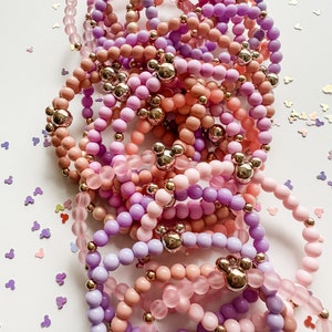 May include: A stack of colorful beaded bracelets with gold Mickey Mouse charms. The bracelets are in shades of pink, purple, and lavender.