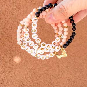 May include: A set of four bracelets with pink and black beads. The bracelets spell out "MAGIC" and "MAMA".