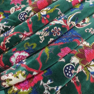 May include: Close-up of a folded, emerald green velvet fabric with a floral pattern. The design features large, stylized flowers in shades of pink, red, and blue, with white and green foliage. The fabric has a soft, luxurious appearance.
