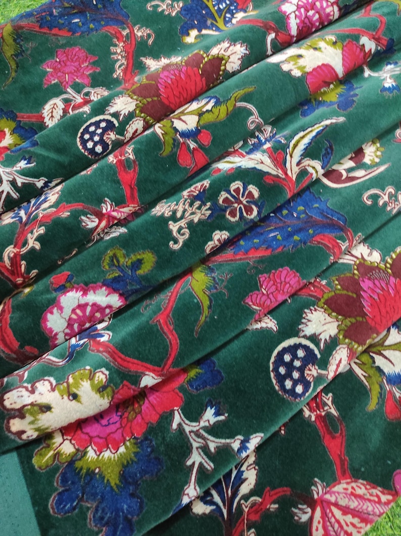 May include: Close-up of a folded, emerald green velvet fabric with a floral pattern. The design features large, stylized flowers in shades of pink, red, and blue, with white and green foliage. The fabric has a soft, luxurious appearance.