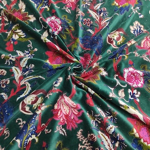 May include: Emerald green velvet fabric with a floral pattern. The design features large pink, red, blue, and white flowers and foliage. The fabric is draped, showing the texture and color variations.