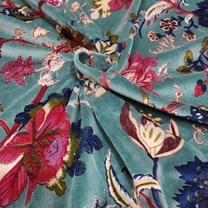 Puede incluir: Tela de terciopelo color turquesa con un estampado floral. El dise&ntilde;o presenta grandes flores estilizadas en tonos rojos, rosas y azules, con hojas y tallos verdes. La tela est&aacute; drapeada, mostrando la textura y profundidad del material.