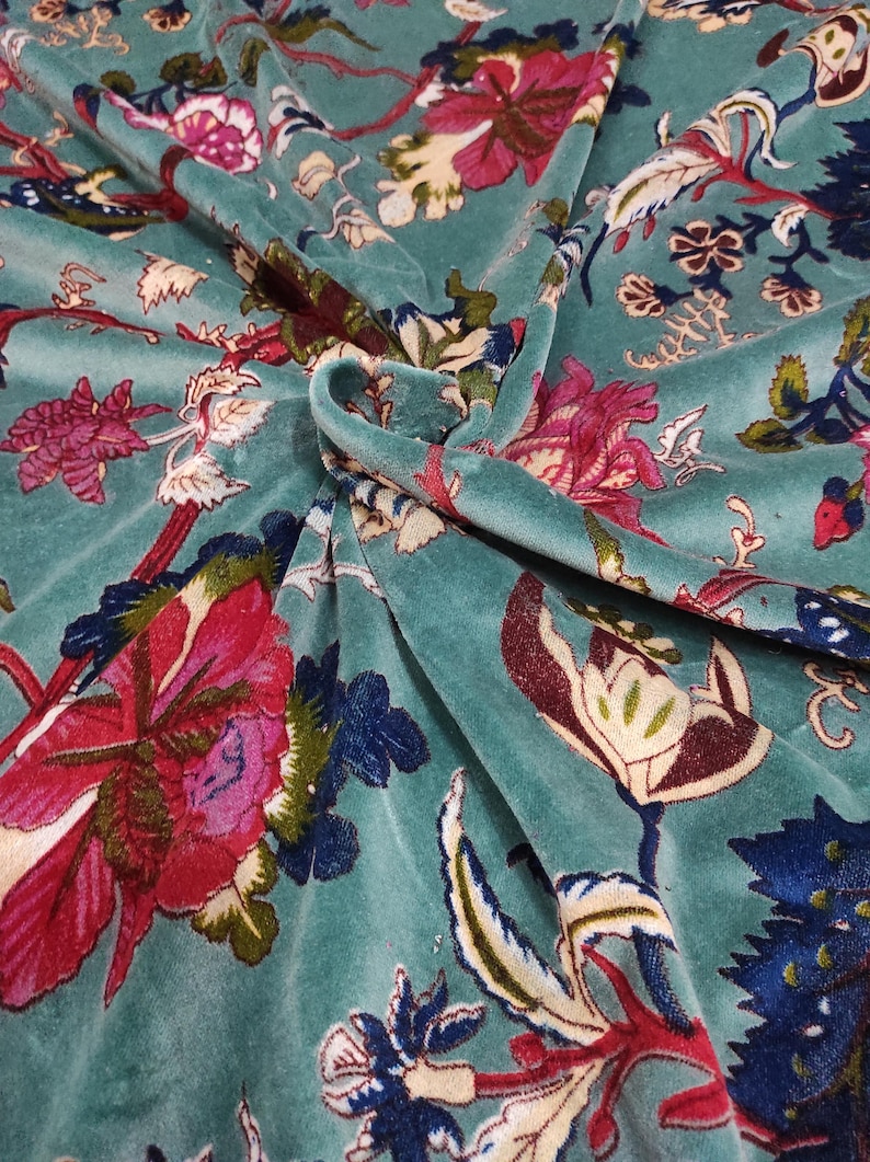 Puede incluir: Tela de terciopelo color turquesa con un estampado floral. El dise&ntilde;o presenta grandes flores estilizadas en tonos rojos, rosas y azules, con hojas y tallos verdes. La tela est&aacute; drapeada, mostrando la textura y profundidad del material.