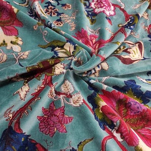 Puede incluir: Tela de terciopelo color turquesa con un estampado floral. El dise&ntilde;o presenta grandes flores estilizadas en tonos rosa, rojo y azul, sobre un fondo turquesa. La tela tiene un aspecto suave y lujoso, con pliegues y textura visibles.