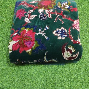 May include: A folded, dark green fabric with a floral pattern. The design features large, vibrant flowers in shades of red, pink, and white, with accents of blue and green. The fabric is placed on a green surface.