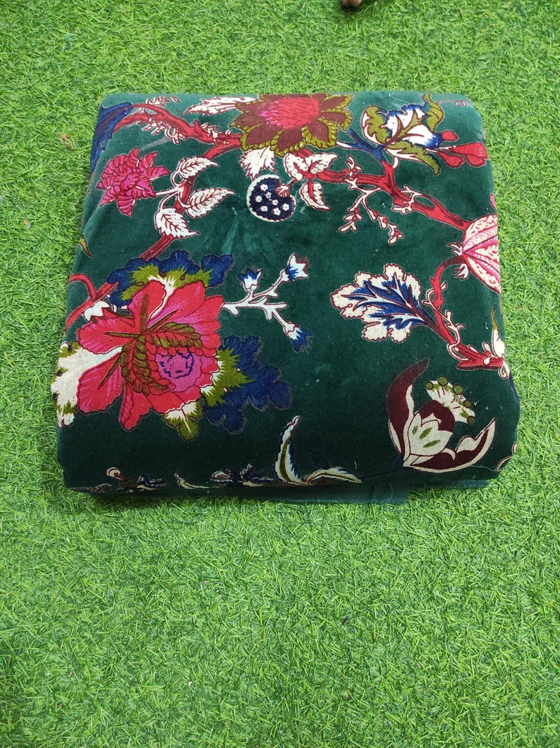 May include: A folded, dark green fabric with a floral pattern. The design features large, vibrant flowers in shades of red, pink, and white, with accents of blue and green. The fabric is placed on a green surface.