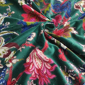 May include: Close-up of a dark green velvet fabric with a floral pattern. The design features large flowers in shades of pink, red, and white, with blue and green leaves and stems. The fabric is folded, showing its texture.