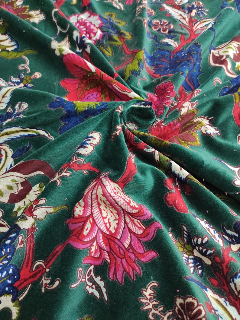 May include: Close-up of a dark green velvet fabric with a floral pattern. The design features large flowers in shades of pink, red, and white, with blue and green leaves and stems. The fabric is folded, showing its texture.