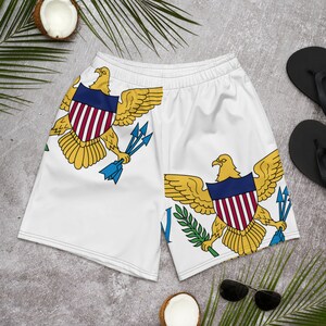 May include: White swimming trunks with a repeated image of the Virgin Islands flag. The flag features a yellow eagle with a blue shield and red and white stripes, three blue spears, and a green branch.