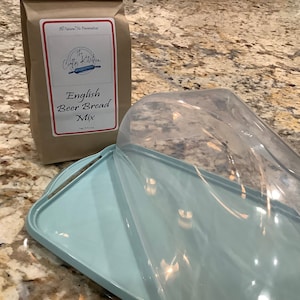 May include: A brown paper bag with a white label that reads "The Crafty Kitchen All Natural No Preservatives English Beer Bread Mix". The bag is sitting on a light brown countertop next to a light blue bread dome.