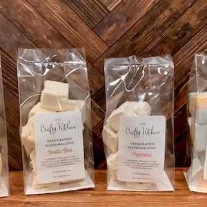 Puede incluir: Cuatro bolsas transparentes de celofán llenas de malvaviscos artesanales. Cada bolsa tiene una etiqueta con el logotipo "The Crafty Kitchen" y los nombres de los sabores: Caramel Bliss, Vanilla Bean, Peppermint y Unicorn. Los malvaviscos son de varios colores y formas.