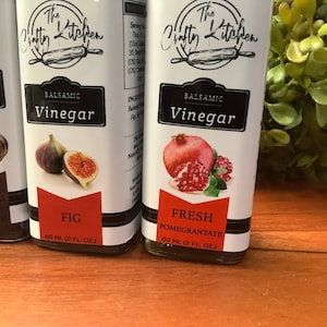 May include: Three glass bottles of balsamic vinegar with white labels. The labels feature a black and white illustration of a rolling pin and the text "The Crafty Kitchen". Each bottle has a different flavor: Traditional, Fig, and Fresh Pomegranate. Each bottle contains 60 ml (2 fl oz) of vinegar.
