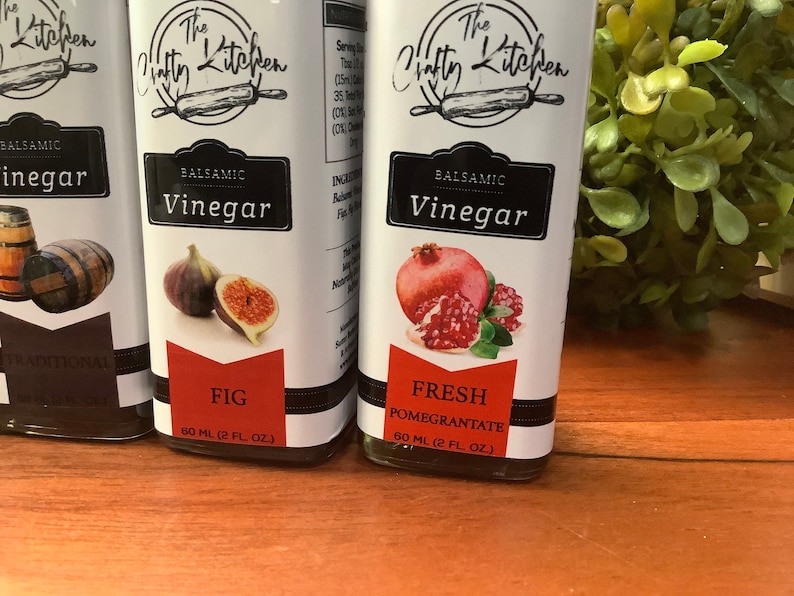 May include: Three glass bottles of balsamic vinegar with white labels. The labels feature a black and white illustration of a rolling pin and the text "The Crafty Kitchen". Each bottle has a different flavor: Traditional, Fig, and Fresh Pomegranate. Each bottle contains 60 ml (2 fl oz) of vinegar.