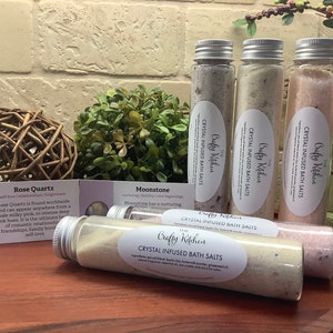 May include: Four glass bottles filled with bath salts, labeled "The Crafty Kitchen Crystal Infused Bath Salts". The bottles are arranged on a wooden surface with a small card describing Rose Quartz and another card describing Moonstone.