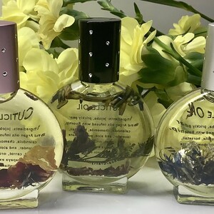 Op de afbeelding: Drie heldere, ronde nagelriemolieflessen met decoratieve doppen. De flessen bevatten gedroogde botanische producten en zijn gelabeld met "Cuticle Oil". De doppen zijn roze, zwart en zilver, elk versierd met kleine strass-steentjes. Gele bloemen staan op de achtergrond.