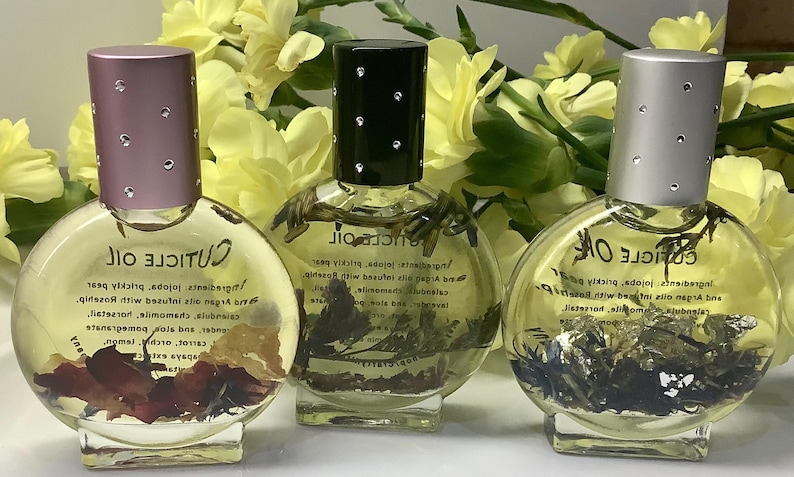 Op de afbeelding: Drie heldere, ronde nagelriemolieflessen met decoratieve doppen. De flessen bevatten gedroogde botanische producten en zijn gelabeld met "Cuticle Oil". De doppen zijn roze, zwart en zilver, elk versierd met kleine strass-steentjes. Gele bloemen staan op de achtergrond.